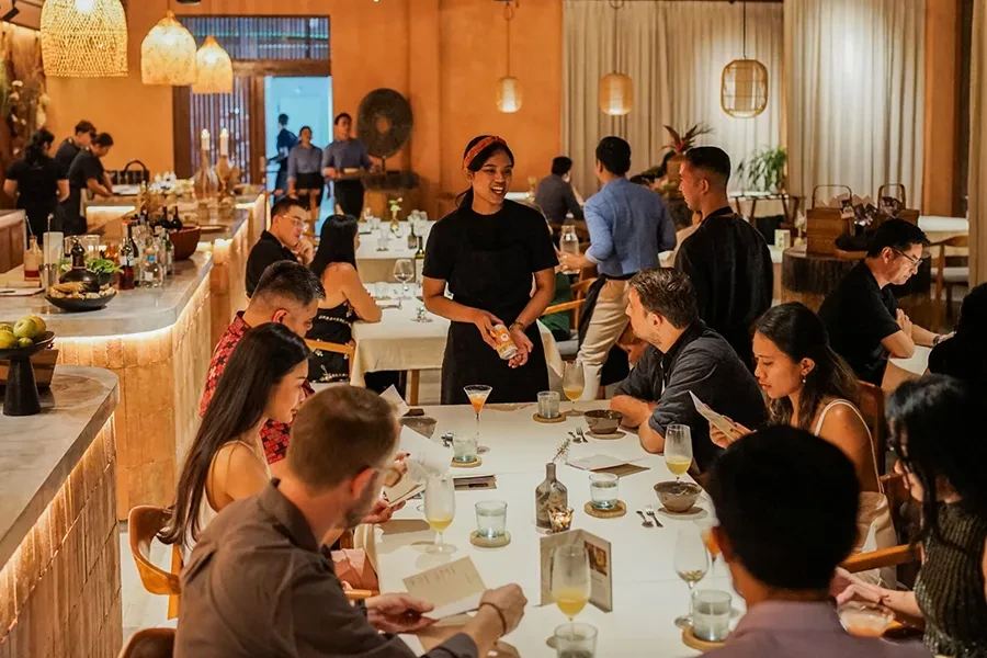An overhead view of the vibrant dining room at Syrco BASE, featuring a large communal table where guests are seated and enjoying drinks. In the background, staff members in black uniforms interact with diners within the warm, earth-toned interior of the restaurant and bar.